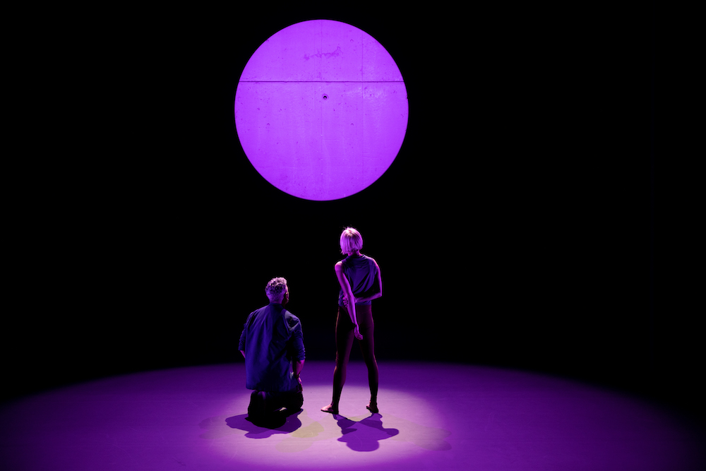 two dancers in circle of light,stare at a purple ball of light on the backwall of their stage space. Their backs are towards us, they are in deep concentration